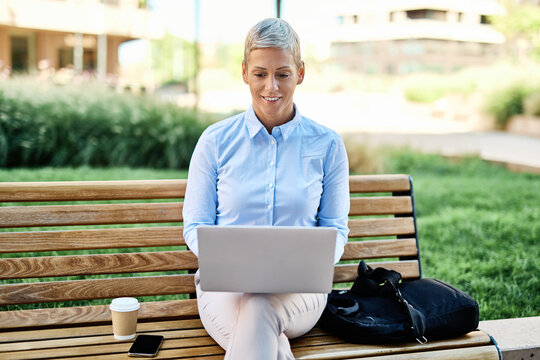 Portrait of a young asian japanese businesswoman woman using a laptop computer and sitting on a bench in a coty park, surrounded by modern corporate office buildings
