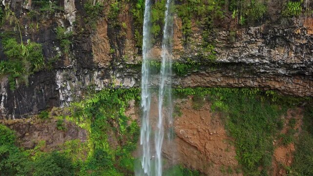 Aerial view shows a tall, narrow waterfall plunging down a rocky cliff. Lush green vegetation blankets the cliff's edges and surrounding terrain. Water cascades in a thin