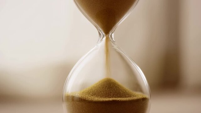 Slow motion close up of hourglass sand flowing and accumulating in lower chamber on soft neutral tabletop