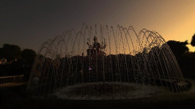 Silhouette of Goddess Durga statue behind water fountain at sunset in India. Dramatic spiritual scene with flowing water, golden sky, and temple ambiance, Chaar Dham Temple in Vrindavan, Uttar Pradesh