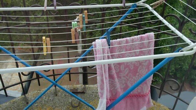 A pink towel hanging on a folding drying rack on the balcony flutters in the wind and slides along the rack. A scene of laundry drying outdoors with clothes pegs, the facade of a residential building,