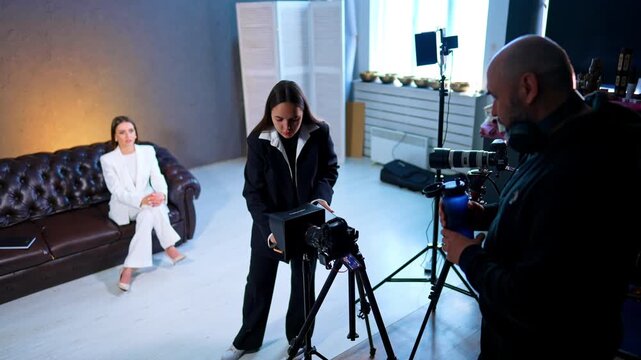 People working behind the scenes arrange filming equipment. Caucasian lady in white suit sits on the sofa waiting for the footage.