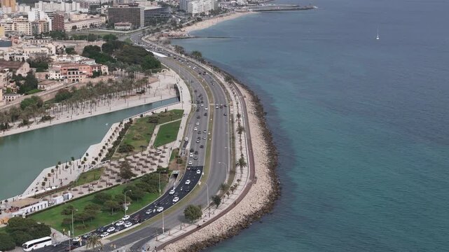 Aerial drone footage of Palma de Mallorca coastal highway and Parc de la Mar lagoon along Mediterranean Sea, Balearic Islands, Spain. Scenic waterfront road with traffic, palm-lined promenade, and tur