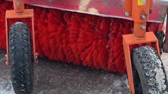 Rotating red cylindrical brushes of a street sweeper machine clean the asphalt road surface during the cold winter season.