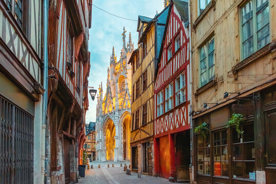 Narrow medieval street in Rouen with half timbered houses leading to illuminated Gothic cathedral facade at sunrise, Normandy, France. Historic architecture and European travel destination