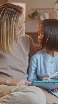 Mother and young daughter sitting on sofa using tablet together in cozy home interior. Parent helping child with learning, drawing or interactive app, or spending quality family time with digital