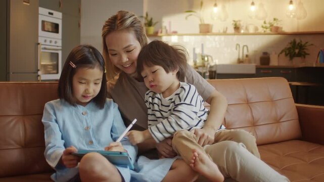Asian mother spending time with two children using digital tablet on sofa at home. Family bonding, online learning and modern parenting lifestyle with kids.