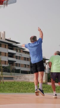 vertical Two senior men playing basketball on an outdoor court, staying active in retirement as one dribbles past his friend and shoots the ball toward the hoop in a park setting