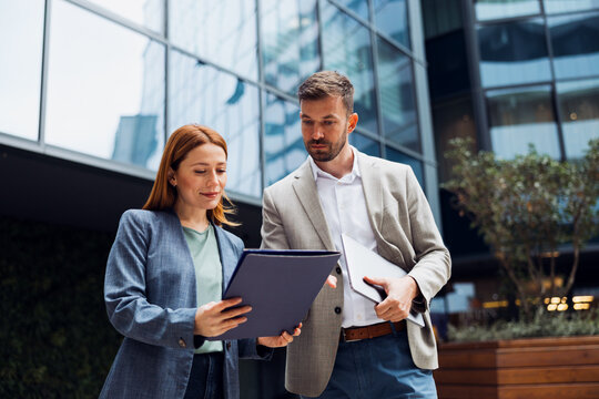 Businesswoman showing a document to her male colleague in front of a modern office building.
