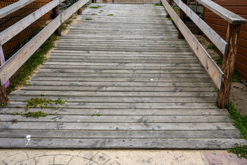 Fototapeta premium a weathered grey wooden boardwalk ramp with railings, featuring natural wood textures and small plants growing through the slats, captured under bright daylight.