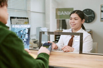Young adult bank teller assisting unseen customer with payment transaction at branch counter,...