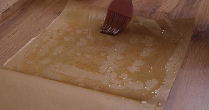 Close-up view of a pastry chef using a silicone brush to grease a sheet of parchment paper with melted butter, preparing for a baking recipe on a wooden kitchen counter.