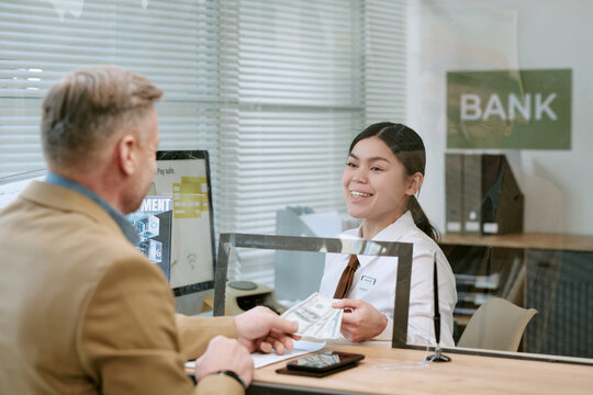 Caucasian middle aged man handing cash to young adult bank teller behind counter, both interacting at branch, woman smiling while processing transaction, glass partition visible