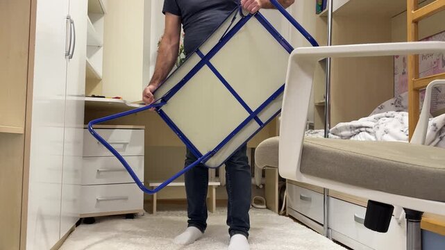 Middle-aged man in blue t-shirt unfolds and assembles a folding table with blue legs in child&rsquo;s room. Practical DIY, home organization, and hands-on lifestyle concept.