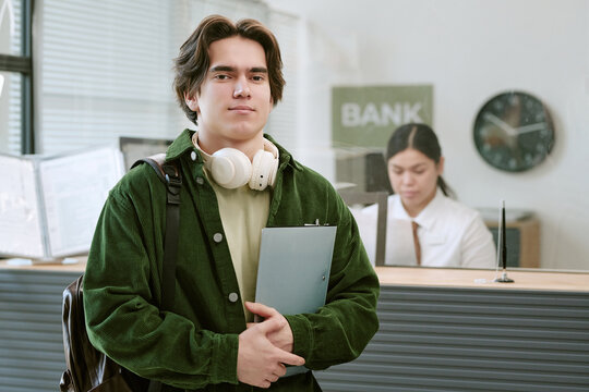 Portrait of Caucasian man standing in modern bank branch, holding folder and headphones around neck while female Asian bank employee working at counter in background