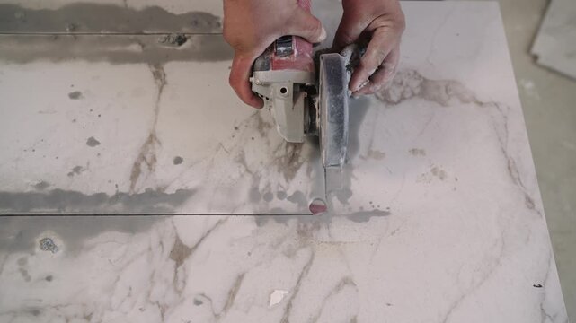 A worker uses an angle grinder with a diamond blade to cut a rectangular hole in a large marble-patterned porcelain tile. Close-up on hands and tool during home renovation or construction.