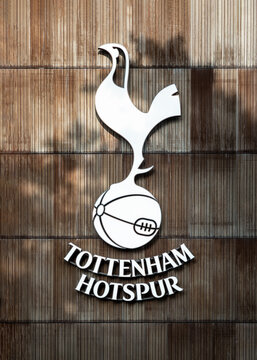 London, UK - Jun 09, 2025 - Dappled sunlight casts soft shadows across the iconic Tottenham Hotspur crest, which is mounted against a textured backdrop of vertical wooden planks. The emblem&rsquo;s sleek, c