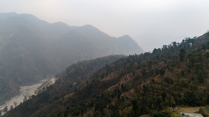 Fototapeta premium Aerial view of Dwara village near Dehradun, Uttarakhand, with a dirt path along a mountain ridge, misty hills, terrace farms, sal and pine forests, and the Song River below on a winter evening.