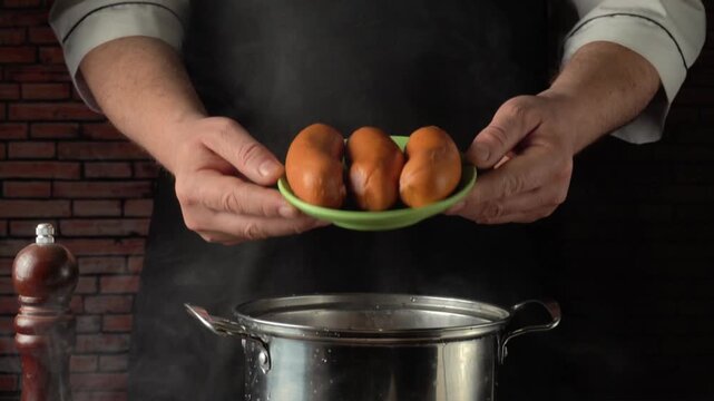Chef holding green plate with boiled sausages over hot pan, demonstrating breakfast preparation process in kitchen with brick wall and wooden pepper mill in foreground