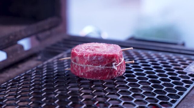 Close-up of beef filet mignon medallion cooking on a charcoal grill with hot embers, BBQ meat preparation outdoors. Juicy steak grilling over fire