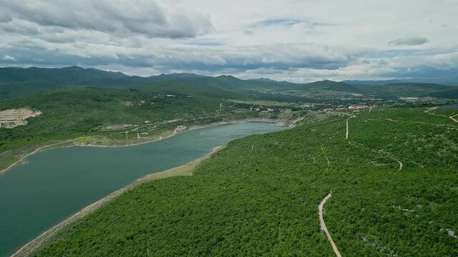 Bosnia and Herzegovina Vlahinja - 05 29 2025 Drone video of turquoise Bileća Lake, green hills, Torič village, Henkel site, quarry, and winding road under a cloudy sky.