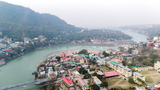 Aerial view of Lakshman Jhula in Rishikesh, Uttarakhand, with the Ganga River flowing below, and temples, houses, and buildings along the riverbank against misty mountains on a winter morning.