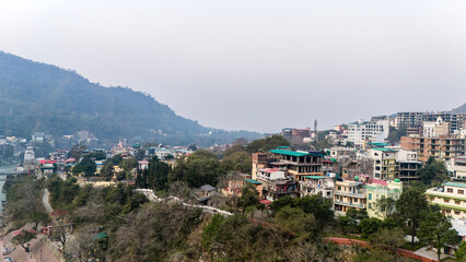 Obraz premium Aerial view of the calm Ganga River in Rishikesh, Uttarakhand on a winter morning, with misty mountains in the background and temples along the riverbank. 