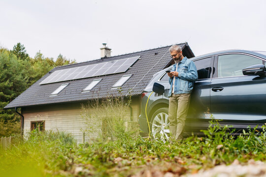 Charging electric car at home, using app. Solar panels on roof behind him.