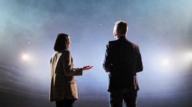 Two speakers standing back to back, engaging in a lively discussion on an empty stage, surrounded by bright spotlights, creating an atmosphere of anticipation before the performance