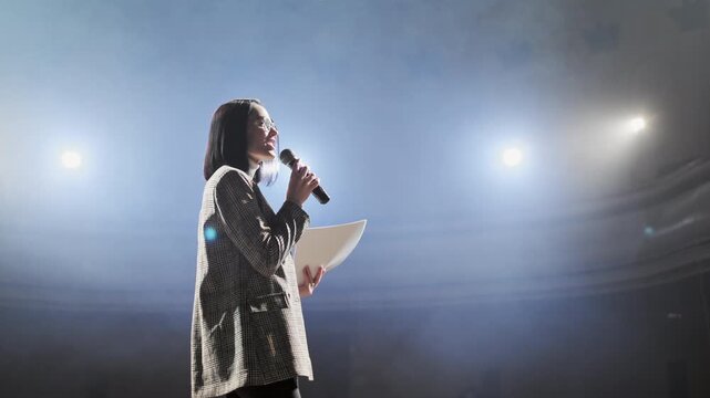 Confident businesswoman in glasses speaking into microphone while addressing audience on stage. Female presenter delivering speech at conference, seminar or corporate event under professional lighting