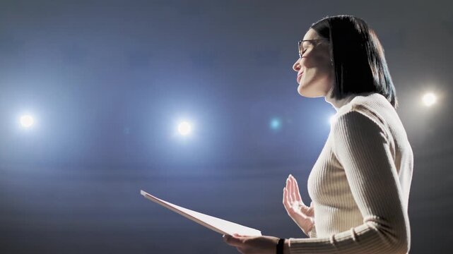 Young woman wearing eyeglasses rehearses her monologue on an empty stage, holding a script under a focused spotlight. She prepares for an upcoming theater performance, highlighting concentration