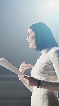 Side view of professional business woman holding notes and speaking on stage under bright spotlight. Female presenter gesturing during speech in corporate or educational setting. Concept of leadership