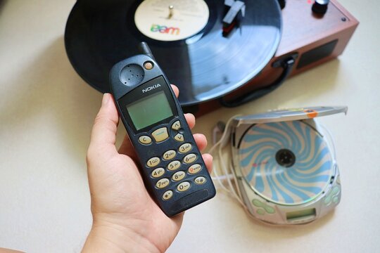 Salvador, Brazil - March 21, 2026: A hand holding a classic Nokia 5120 mobile phone from the late 1990s next to a portable CD (discman) player and a vintage record player turntable.