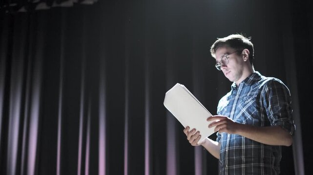 Theater director standing on a dark stage, illuminated by a spotlight, using a tablet to guide a rehearsal. Pointing at the screen with precision, he directs the performance with focus and authority
