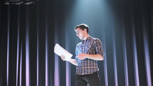 Focused young man standing on stage under bright spotlight holding script and gesturing while speaking. Actor rehearsing lines or practicing performance in theater setting with dramatic lighting and