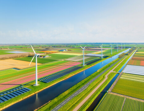 Aerial view of Dutch countryside with wind turbines and solar panels among green fields. Sustainable energy and agriculture in Netherlands under blue sky. Summer sunny day rural landscape from above.