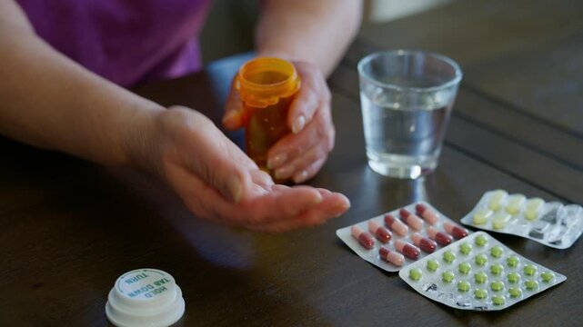 Close up of elderly hands pouring pills from an orange prescription bottle into the palm with blister packs of medication on the table.