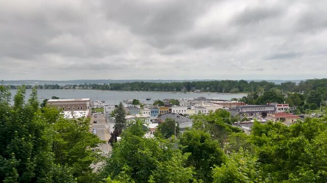 High-angle shot from a hillside bluff overlooking Harbor Springs, MI. Lush greenery leads down to the historic downtown, marina, and tranquil Little Traverse Bay on a moody day.