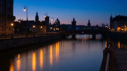 Fototapeta premium Golden City Lights Reflecting on River at Blue Hour Twilight. Warm amber streetlights shimmer across calm water surface during magical blue hour, urban skyline silhouette in soft focus background