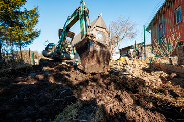 Fototapeta premium Compact excavator digs a muddy trench in a fenced yard beside two houses, with a dog near soil and tools. Low angle, sharp sunlight, long shadows, clear daytime scene.