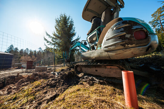 A compact tracked excavator works beside a wire fence and tall spruce, with a worker in the cab at midday. Fresh soil, orange drainage pipe, and rocks suggest trenching.