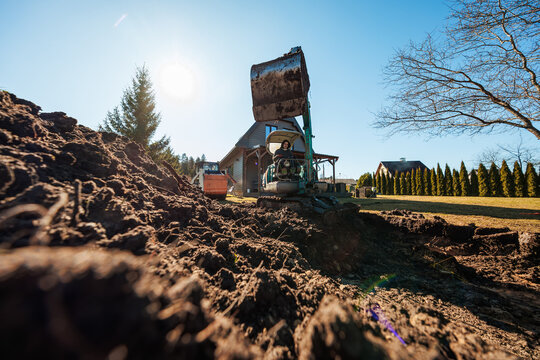 A mini excavator with a mud caked bucket digs beside a modern wood sided house on a large lawn, piles of fresh soil and evergreens present under bright afternoon sun.
