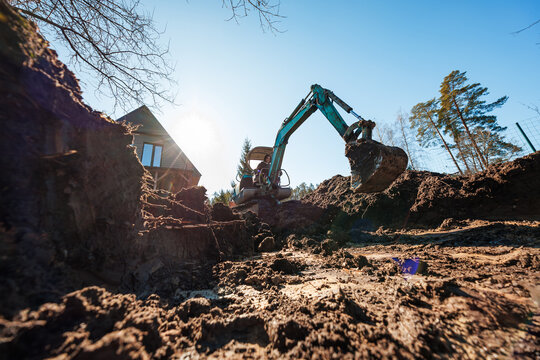 A compact teal excavator digs a wide trench by a modern A frame cabin at dawn, bucket raised with wet soil in backlit morning sun on a forested residential lot.