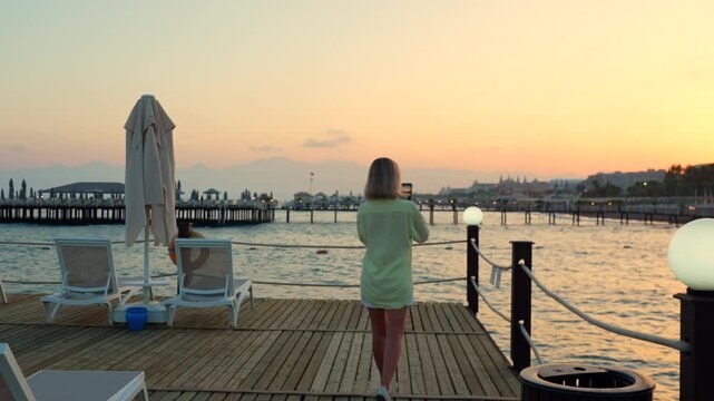 Woman walks on pier and records sea on phone at sunset