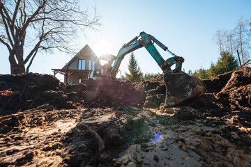 Fototapeta premium A compact excavator digs a wide trench near a modern A frame cabin as low sun flares, long shadows stretch, and conifer trees and a wire fence frame the rural work site.