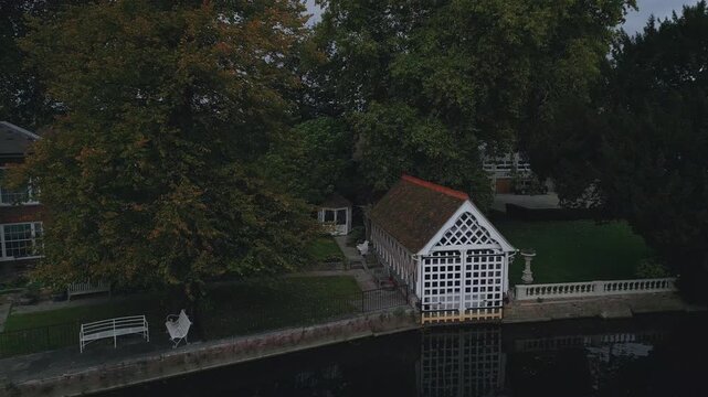 A charming boathouse with a white latticed facade stands on a canal, reflected in the dark water. Mature trees provide lush shade.