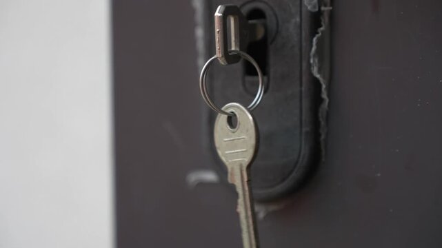 Extreme close-up (ECU) focusing on a silver key hanging and swinging in a door lock. Detailed view of the key's teeth and the metallic texture of the lock cylinder. Professional 4K footage at 25fps 