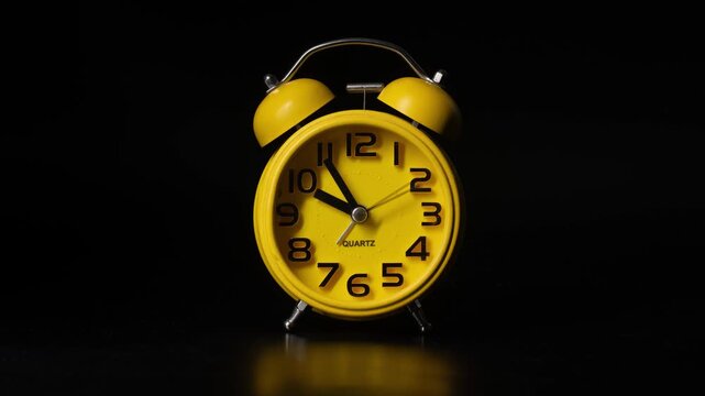 Close up timelapse video of a yellow bedside alarm clock showing the hands moving rapidly against a black background.