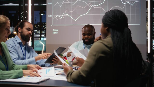 Diverse professionals group evaluating files and performance indicators, using projections and diagrams to align the company objectives with investors expectations for company progress. Camera A.