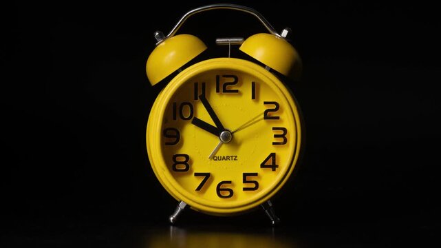 Close up timelapse video of a yellow bedside alarm clock showing the hands moving rapidly against a black background.